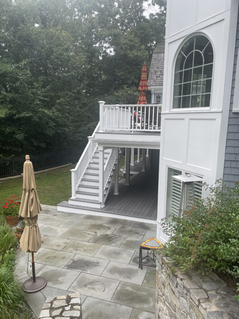 White outdoor staircase leading to a deck above a stone patio surrounded by greenery.